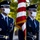 Two U.S. Air Force honor guard members stand at attention in dress blue uniforms while wearing white face masks, holding ceremonial flags including the American flag. The personnel display rank insignia, service ribbons, and ceremonial accessories consistent with a formal color guard presentation. The outdoor setting features blurred green foliage in the background, suggesting a daytime ceremonial event.