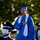 A man wearing a blue graduation cap and gown with a white stole stands with his right hand placed over his heart, likely during a national anthem or pledge. In the foreground, uniformed personnel wearing face masks and white gloves are visible, suggesting a formal military or honor guard ceremony. The setting appears to be an outdoor graduation event with trees visible in the background.