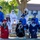 A group of approximately 12 graduates are seated on bleachers or wooden benches outdoors, wearing academic caps and gowns in various colors including blue, white, red, black, and green. All individuals are wearing face masks, suggesting this ceremony took place during the COVID-19 pandemic period. The setting appears to be a school or institutional campus with trees and a building visible in the background.