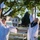 Two young women in white graduation gowns are celebrating outdoors, tossing their mortarboard caps into the air. One graduate is also wearing a navy blue stole with gold trim, suggesting academic distinction. The scene takes place in what appears to be a school or campus area with trees, buildings, and bleachers visible in the background.