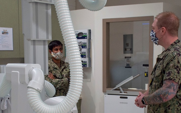 Four U.S. Navy personnel in NWU Type III (digital woodland camouflage) uniforms are gathered in what appears to be a medical radiology or X-ray room. All individuals are wearing face masks, suggesting this was taken during the COVID-19 pandemic period. One service member appears to be briefing or demonstrating equipment to the others.