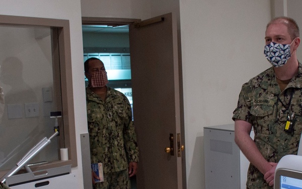 Three U.S. Navy personnel in camouflage uniforms (NWU Type III) are gathered in what appears to be a medical facility room. All three individuals are wearing face masks, consistent with COVID-19 precautions. The scene suggests a briefing or inspection near medical equipment, including what appears to be a digital medical device with a touchscreen interface.