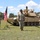 1st Battalion, 8th Cavalry Regiment Change of Command Three U.S. Army soldiers in MultiCam/OCP uniforms are participating in a formal military ceremony outdoors, with an American flag on a stand and an M1 Abrams main battle tank in the background. Two of the soldiers are wearing black cavalry-style campaign hats, suggesting affiliation with a cavalry unit. The setting appears to be a military training area or installation, likely in Europe based on the landscape.