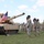 1st Battalion, 8th Cavalry Regiment Change of Command A U.S. Army military ceremony is taking place outdoors on a grass field, featuring soldiers in formation saluting alongside an M1 Abrams main battle tank displaying an American flag. The soldiers are wearing distinctive black cavalry Stetson hats, suggesting this is a unit associated with U.S. Army Cavalry. A speaker system on a tripod stand is visible to the right, indicating a formal ceremonial event.
