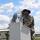 1st Battalion, 8th Cavalry Regiment Change of Command A U.S. Army soldier wearing a black cavalry Stetson hat and face covering stands at a white outdoor podium during what appears to be a military ceremony or formation. A clear plexiglass shield and a hand sanitizer bottle are visible on the podium, indicating COVID-19 safety protocols. Additional soldiers in similar uniforms are visible in the background near military-style buildings.