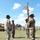 1st Battalion, 8th Cavalry Regiment Change of Command A U.S. Army military ceremony, likely a change of command or activation ceremony, is taking place on an outdoor parade field. Several soldiers in OCP (Operational Camouflage Pattern) uniforms and cavalry-style campaign hats are participating, with one soldier holding a unit guidon adorned with multiple campaign streamers. An M1 Abrams tank is visible in the background left, and additional soldiers are seated and standing in formation.