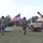 1st Battalion, 8th Cavalry Regiment Change of Command Three U.S. Army soldiers in MultiCam (OCP) uniforms and black cavalry Stetson hats walk toward armored vehicles on a grassy field. A U.S. flag on a portable stand is positioned between the vehicles. The scene appears to be a military ceremony or inspection at what looks like a training area or base.