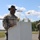1st Battalion, 8th Cavalry Regiment Change of Command A U.S. Army soldier wearing a black cavalry hat and face covering stands at an outdoor podium with a microphone, appearing to address an audience at a military ceremony. A plexiglass shield and hand sanitizer dispenser are visible on the podium, indicating COVID-19 safety protocols. A second soldier in similar attire stands in the background.