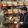Three individuals wearing face masks are conducting an inventory or inspection activity in a military or government warehouse facility. A person in U.S. Navy digital camouflage (NWU) uniform is holding a small packaged item and showing it to two civilians, one of whom is holding a clipboard. The shelving units are orange industrial racks with numerous cardboard boxes labeled with barcodes and bin location numbers.