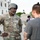 A U.S. Army soldier identified by name tape as 'Matthews,' wearing OCP camouflage uniform and a combat helmet, is engaged in a conversation with a civilian male who has visible tattoos and a man-bun hairstyle. The soldier displays a 2nd Infantry Division patch on his left shoulder and Staff Sergeant rank insignia. The setting appears to be near a government building with security fencing visible in the background.