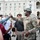 A U.S. Army soldier in camouflage uniform and tactical helmet is engaged in a fist-bump gesture with a civilian in front of a large neoclassical government building. A crowd of civilians surrounds the scene, including a photographer with a professional camera. Several individuals are wearing face coverings, suggesting this may be during a public gathering or protest event.