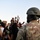 A large outdoor protest is depicted with a crowd of demonstrators facing a person in military camouflage uniform and helmet in the foreground. Protesters are holding signs, raising fists, and using a megaphone, while photographers document the event. A visible sign references George Floyd, indicating this is a racial justice protest.