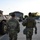 Two soldiers in U.S. Army MultiCam (OCP) camouflage uniforms are walking away from the camera toward a crowd gathered near a neoclassical building. An armored military vehicle, consistent with an M1117 Guardian or similar wheeled armored personnel carrier, is parked to their left. A temporary chain-link security fence is visible on the right side, suggesting a security or crowd-control deployment.