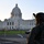 A military or National Guard soldier in camouflage uniform and combat helmet stands in the foreground, facing a large white domed government building. The scene appears to be taken at dusk or dawn, with the soldier positioned as if on guard duty. The building in the background is a classical neoclassical-style capitol structure.