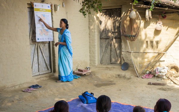 A woman in a blue sari is conducting an outdoor community education session in what appears to be a rural South Asian village. She is pointing to an informational poster mounted on a mud-brick wall while a group of approximately 15 women sit on a blue mat on the ground facing her. The setting suggests a grassroots health or community awareness program.