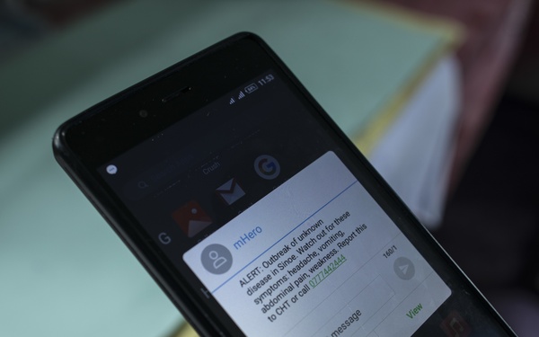 A person is holding an Android smartphone displaying an mHero health alert notification about an outbreak of unknown disease in Sinoe. The message warns about symptoms including headache, vomiting, abdominal pain, and weakness, and instructs recipients to report to CHT or call a provided phone number. This appears to be a public health emergency communication system in use, likely in a West African context.