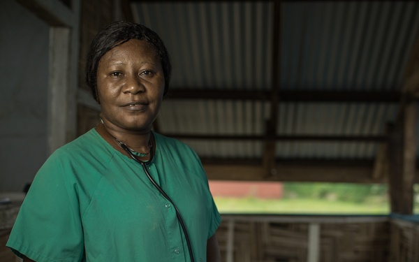 A female healthcare worker wearing green medical scrubs and a stethoscope around her neck stands in what appears to be a rural or basic medical facility. She is posed facing the camera with a calm, composed expression. The setting suggests a low-resource healthcare environment, likely in a sub-Saharan African context based on the construction materials and surroundings.