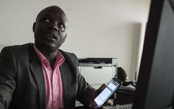 A man in a dark blazer and pink shirt sits at an office desk, holding a smartphone with an active screen while also using a computer mouse. He appears to be multitasking, looking upward possibly in thought or conversation. The office setting includes a desktop computer monitor, keyboard, printer, and additional devices on the desk.