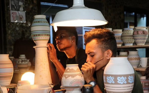 A pottery workshop scene showing multiple artisans decorating ceramic vessels under bright workshop lighting. Several young men are engaged in hand-painting and detailing clay pots and vases with geometric and traditional patterns. The workspace is densely populated with tools, pigment containers, brushes, and finished and unfinished ceramic pieces.