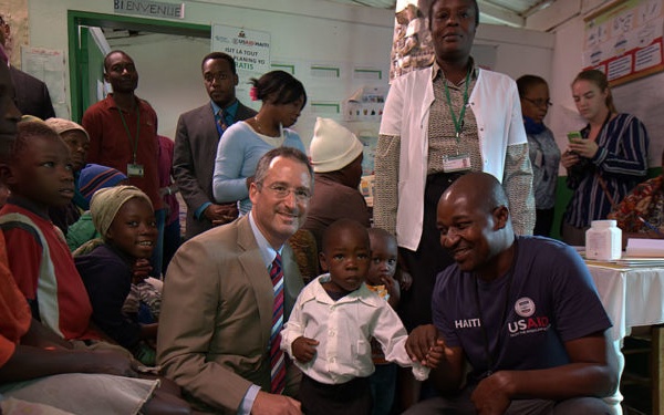The image shows a group of people gathered inside what appears to be a health clinic or community center in Haiti. Two men are crouching down posing with a young toddler in the foreground, one wearing a suit and tie and the other wearing a USAID Haiti t-shirt. The setting includes medical staff, community members, and children in a crowded indoor facility.
