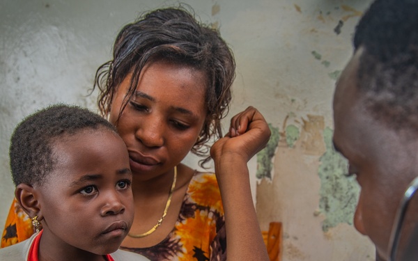A healthcare worker is examining a young child using a stethoscope while a woman, likely the child's mother, holds the child and looks on attentively. The examination appears to be taking place in a modest medical facility with worn walls. The child appears alert but apprehensive during the examination.