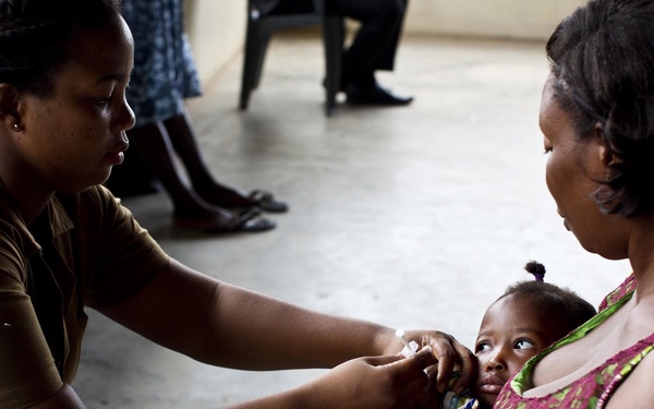 A healthcare worker in a tan/khaki uniform is administering oral drops or medication to a young infant being held by its mother. The setting appears to be a clinic or health facility, likely in sub-Saharan Africa based on the clothing and environment. This appears to be a routine immunization or oral vaccination procedure.