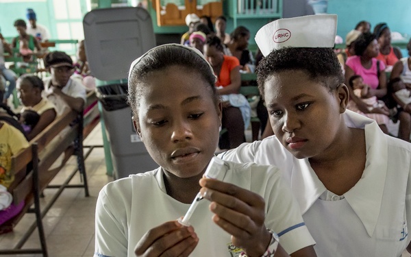 Two nurses in white uniforms are examining a syringe or medical vial in what appears to be a busy healthcare clinic waiting room. The nurse on the right wears a cap with 'URHC' printed on it, suggesting an institutional affiliation. The background shows numerous patients, predominantly women and children, seated on wooden benches waiting for care.