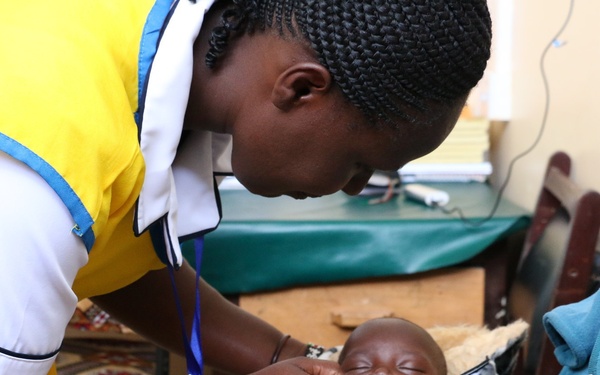 A healthcare worker in a yellow and white uniform is administering oral medication or drops to a swaddled infant being held by a woman, likely the mother. The setting appears to be a clinical or community health facility. The scene suggests a routine immunization or medication administration procedure.
