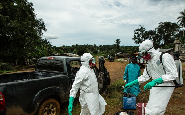 The image shows health workers in full personal protective equipment (PPE) including white hazmat suits, respirator masks, and green gloves, operating in a rural African village setting. A worker wearing a backpack sprayer appears to be decontaminating or preparing equipment near a pickup truck. A sign reading 'GOOD BYE ARTHINGTON' is visible in the background, suggesting this is near the town of Arthington, Liberia, likely during the 2014-2016 Ebola outbreak response.