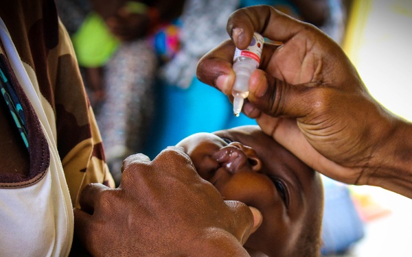 A healthcare worker is administering oral drops to a young infant being held by an adult, likely a parent or caregiver. The small vial with a dropper tip is being directed into the baby's open mouth, consistent with oral vaccine administration such as a polio vaccine. The setting appears to be an outdoor or semi-outdoor health clinic in what appears to be a sub-Saharan African context.