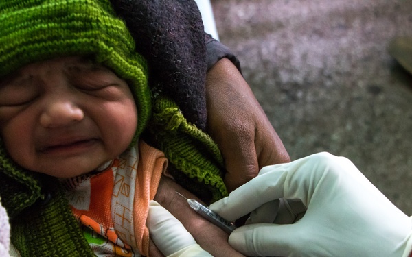 A young infant wearing a green knit hat is receiving an injection in the arm, likely a vaccination. The baby appears to be crying or distressed during the procedure. A healthcare worker wearing white latex gloves is administering the shot while an adult holds the child.