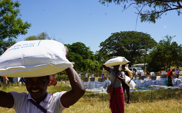 A smiling young person carries a white bag labeled 'Maize Meal 20 kg Net' on their head in an outdoor setting. Behind them, other individuals are similarly carrying bags, with a large distribution area visible in the background containing numerous stacked white sacks. The scene appears to depict a food aid or relief distribution event.