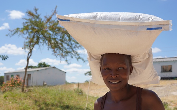 A woman is carrying a large white sack balanced on her head while holding another bag in her hand, smiling at the camera. She is outdoors in what appears to be a rural African village setting with white-painted structures visible in the background. The scene is bright and sunny with scattered clouds and green vegetation.