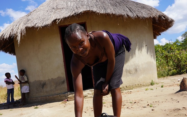 A woman is cooking outdoors over an open fire made from logs and stones, in front of a traditional round mud-walled hut with a thatched roof. The setting appears to be a rural African village with dry, sandy ground and lush vegetation visible in the background. Two children can be seen standing near the entrance of the hut in the background.