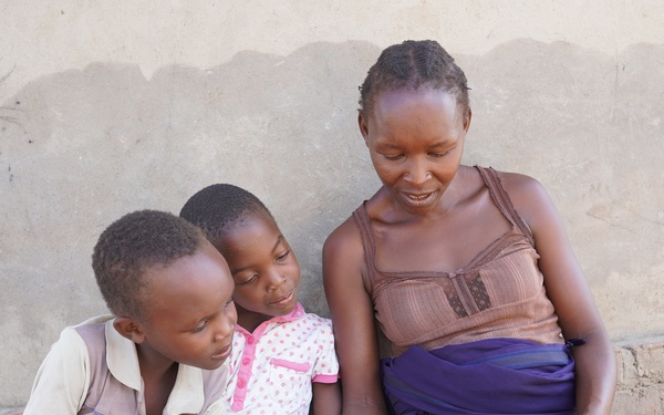 A woman sits outdoors against a plastered wall with two young children, reading from a small book together. The group appears engaged and attentive, with the children leaning in to look at the pages. The setting suggests a modest rural or semi-urban environment.