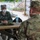 Two military personnel in camouflage uniforms are seated across from each other at an outdoor picnic table, engaged in conversation. The individual facing the camera wears a name tape reading 'ZIMMER' and has a face mask pulled down, while the person opposite wears an Air Force uniform with a face mask properly worn. The setting appears to be a covered outdoor area, suggesting a COVID-era military interaction.