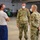 Three military personnel are engaged in a conversation inside a large hangar or warehouse facility. Two individuals are wearing U.S. Army OCP (Operational Camouflage Pattern) uniforms, while the third is in a gray t-shirt and blue pants with a rank insignia visible. All three are wearing face masks, suggesting this was taken during the COVID-19 pandemic period.