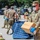 Military personnel in OCP (Operational Camouflage Pattern) uniforms are conducting what appears to be a COVID-19 relief or food distribution operation in an outdoor parking lot setting. All visible personnel are wearing face masks and blue nitrile gloves as protective equipment. The foreground soldier is carrying a cardboard box, consistent with supply distribution activities.