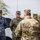 Four military personnel are gathered outdoors in what appears to be a briefing or meeting scenario. Three individuals wearing Romanian military uniforms (including one in blue digital camouflage and two in green woodland camouflage) face a fourth person in OCP/MultiCam pattern uniform whose back is to the camera. Romanian national flag patches are visible on multiple uniforms.