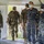 A group of military personnel from multiple nations are gathered inside a large white tent, all wearing surgical face masks. At least two distinct uniform types are visible: Romanian military uniforms (blue-grey digital camouflage with 'ROMANIA' name tape and Romanian flag patch) and US Army OCP (Operational Camouflage Pattern) uniforms. The setting appears to be a multinational military exercise or joint operation event, likely during the COVID-19 pandemic period.