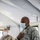 A U.S. Air Force service member wearing the Airman Battle Uniform (ABU) and a blue surgical face mask stands inside what appears to be a military tent or temporary structure, with his right hand placed over his chest. A second military personnel member in a different camouflage pattern (OCP) is visible in the background, also wearing a face mask. The setting and face masks suggest a COVID-19 era military operation or ceremony.