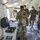 A group of military personnel from multiple nations are gathered inside a military field tent, examining equipment on a table. All individuals are wearing face masks, suggesting this was taken during the COVID-19 pandemic period. The setting appears to be a joint military exercise or briefing involving coalition forces.