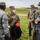 Four military personnel wearing face masks are engaged in a discussion outdoors on a grassy area, likely during a joint military exercise or coordination meeting. The group includes U.S. Air Force members and Romanian military personnel, identifiable by their distinct uniforms and insignia. The setting appears to be near a military installation or airfield.