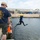 A fully equipped scuba diver in a black wetsuit and yellow helmet is mid-jump off a pier into a harbor, while support personnel observe from the dock. The man in the foreground wears a navy blue t-shirt reading 'Norfolk NADS Tradition Divers,' indicating this is a U.S. Navy diving unit activity. Industrial waterfront buildings and inflatable boats are visible in the background.
