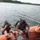Two scuba divers in full diving gear are seated on the edge of an orange inflatable rescue boat, appearing to prepare for or return from a dive. A third person wearing a tan cap, sunglasses, and a military-style t-shirt with a patch stands on the boat observing them. The scene takes place on a calm body of water with a tree-lined shoreline visible in the background.