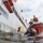 A low water-level perspective shows military or naval personnel conducting what appears to be a hull inspection or underwater demolition/diving operation alongside a large vessel marked 'FR-10-2'. A diver is visible in the water near the ship's hull, while several personnel in military-style clothing observe from an orange rigid inflatable boat (RIB). A yellow device is attached to the ship's hull, suggesting a training or inspection exercise.