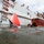 Military or naval personnel are conducting a diving operation alongside a gray vessel marked 'FR-70-2'. Two divers are visible in the water near the hull, while several personnel on the vessel's deck and an orange rigid inflatable boat (RIB) provide support. A large red panel or patch is being positioned against the ship's hull, suggesting a hull repair or inspection exercise.