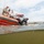 A water-level photograph captures military or coast guard personnel on an orange rigid inflatable boat (RIB) as a scuba diver enters or exits the water from the vessel's side. Several individuals in military uniforms and civilian attire observe from the boat and a larger vessel in the background. The scene appears to take place near a port or naval facility visible on the shoreline.