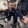 A U.S. Navy sailor named Williams is working on mechanical or aviation equipment aboard what appears to be a naval vessel. He is crouching down and using a screwdriver with an orange handle to work on components held in metal brackets or trays. At least one other Navy personnel is visible standing nearby in the background.