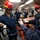 U.S. Navy personnel are conducting a damage control or firefighting training drill aboard a naval vessel. Several sailors wearing red firefighting helmets and navy blue coveralls are gathered around equipment, with an instructor appearing to demonstrate proper use of gear. An orange equipment case is visible on the deck, and the setting is clearly an interior compartment of a warship.